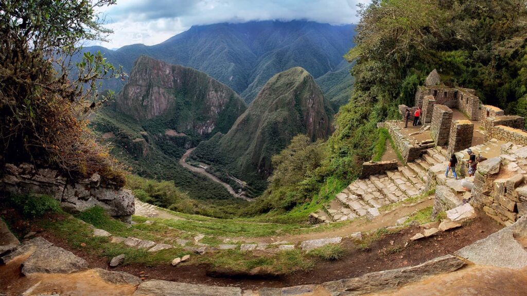 Tourists gazing at the Machu Picchu ruins from the Sun Gate in Cusco, surrounded by lush green mountains and clear skies.